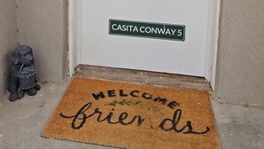 A white door with a welcome wreath, a doormat with 'welcome friends' text, and a street sign reading 'CASITA CONWAY 5'.