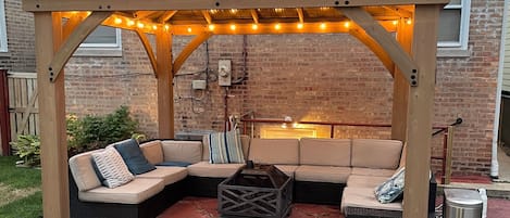 A covered outdoor seating area with a curved sofa and a coffee table, under a metal roof with string lights.