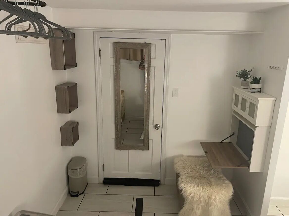 A small, compact room with a white door, a mirror, a trash bin, a fur rug, and a small wooden shelf with plants.