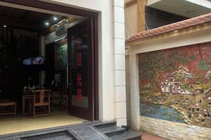 An entrance with a traditional Chinese mural, a wooden door with red characters, and a small table with chairs inside. - HOANG HA VILLA (Bac Ninh)