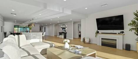 A modern living room with a large flat-screen TV, a fireplace, and a dining area in the background.