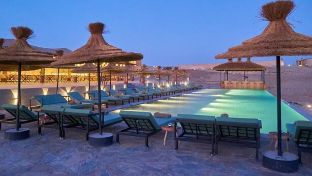 Zona de piscina con sillones de descanso y sombrillas de paja, rodeada por un paisaje desértico.