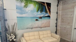 A beige sofa in a room with a beach scene mural, a wooden floor, and a potted plant with white flowers. - Great room w/ pool table in a 1-bdrm party barn in Newaygo; River access (Newaygo)