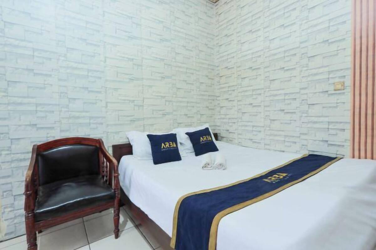 A hotel room with a brick-patterned wall, a wooden chair, a bed with a blue and gold bedspread, and two pillows with a logo.