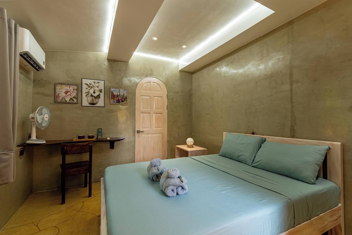 A bedroom with a bed, a desk, a chair, a nightstand, and a wall-mounted air conditioner.