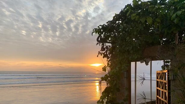 Vista de playa con un atardecer, una estructura de madera con ventanal de celosía y un muro cubierto de hiedra.