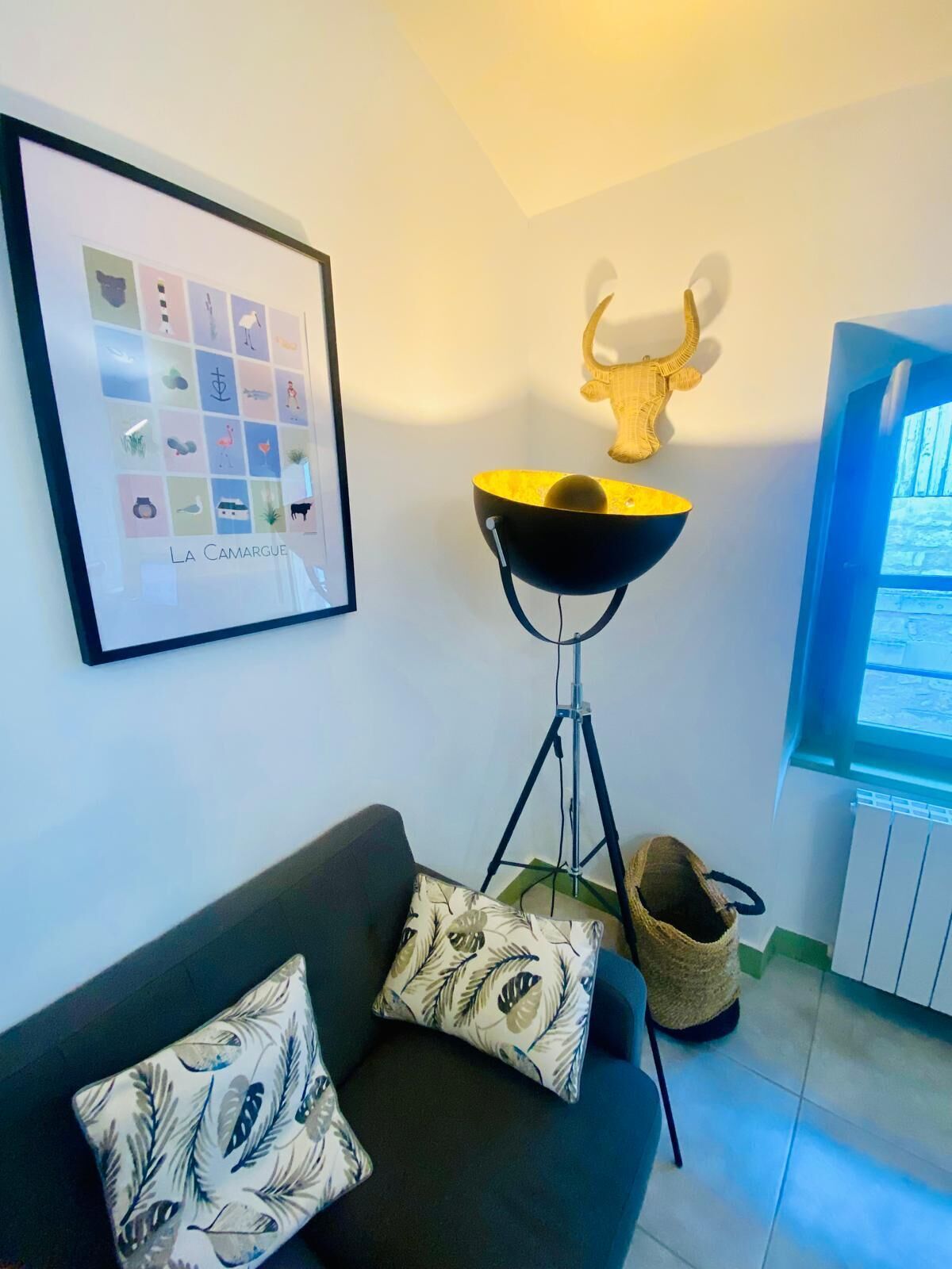 A living room with a black sofa, patterned cushions, a framed artwork, a floor lamp, a decorative wall piece, and a window with blinds.