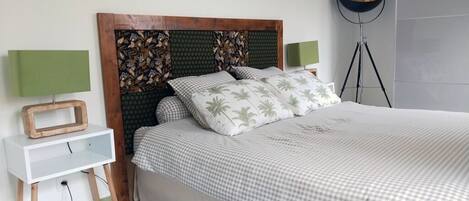 Ein ordentlich bezogenes Bett mit karierter Tagesdecke und Kissen mit Palmenblatt-Muster, ein hölzernes Kopfteil mit Blumenmuster, ein Nachttisch mit grüner Lampe und eine Stehlampe mit goldfarbenem Fuß.