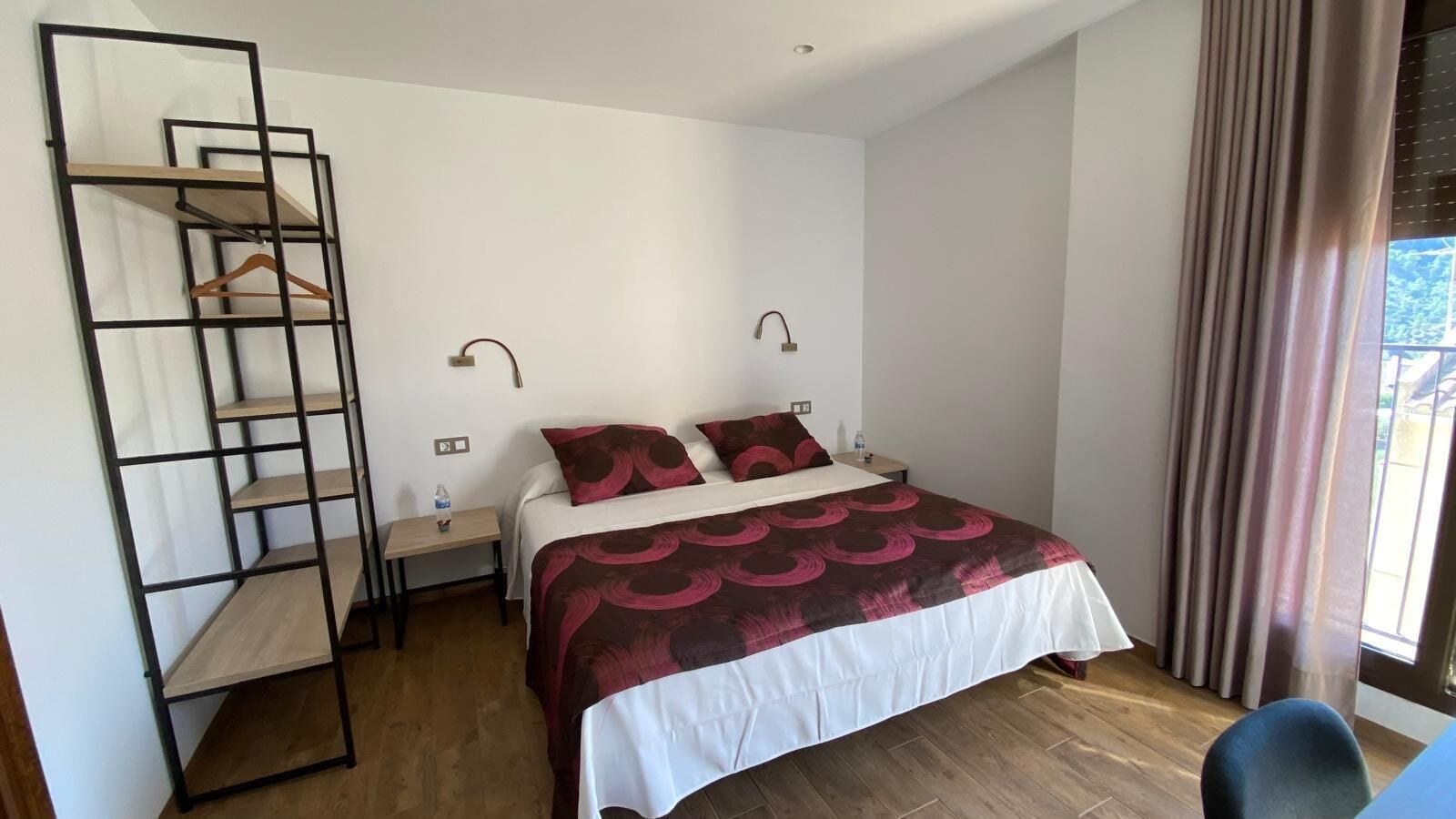 Quarto com cama, chão de madeira, estante de metal, mesa de cabeceira e janela com cortinas.