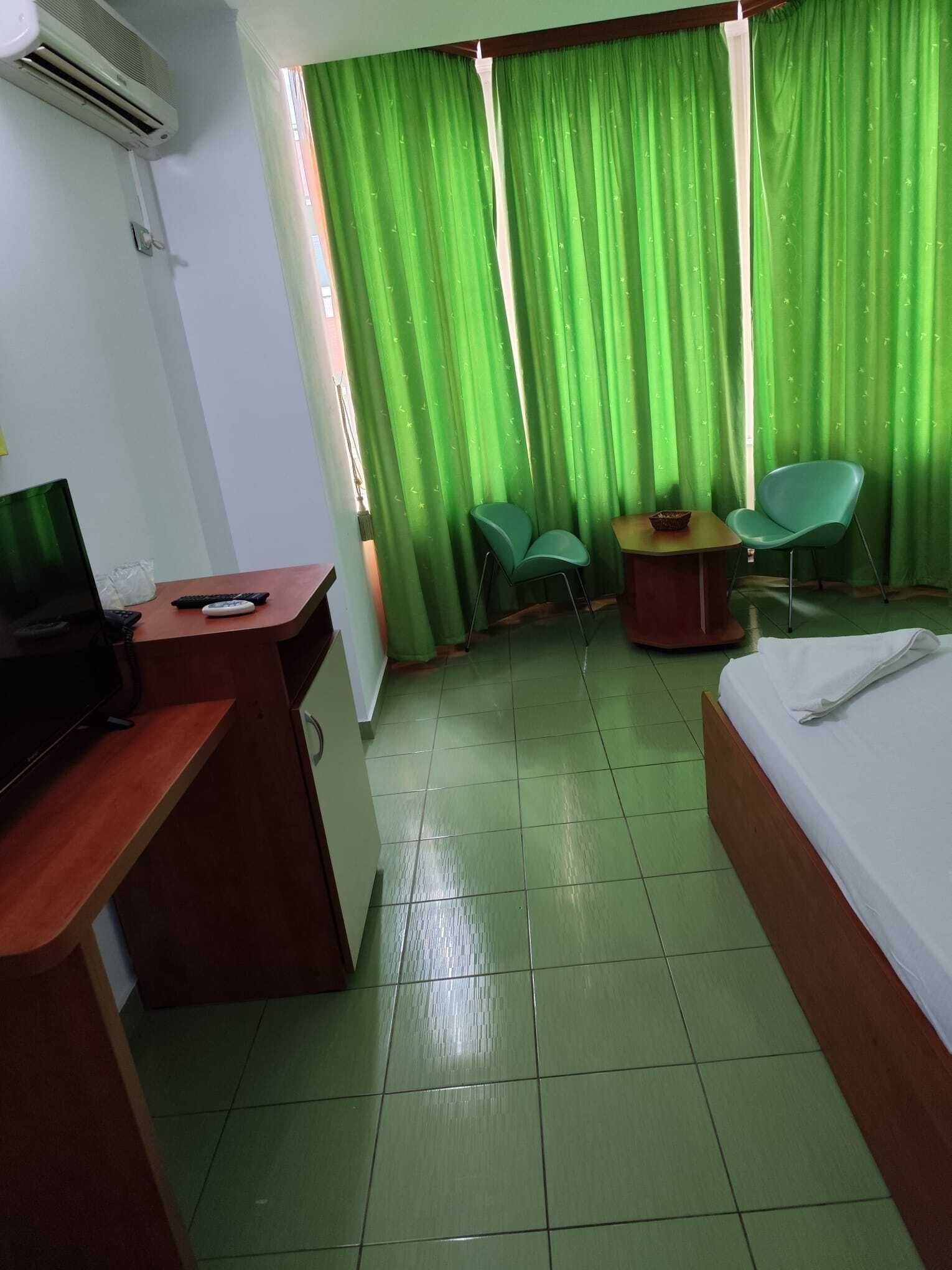 Hotellihuone, jossa on sänky, työpöytä tietokoneella, kaksi tuolia, pieni pöytä ja vihreät verhot.
