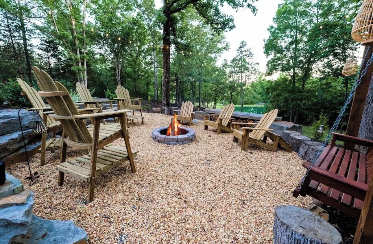 Un área de estar al aire libre con sillas y mesas de madera, un brasero y un paisaje boscoso natural.