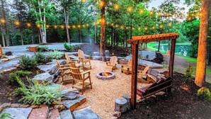 An outdoor seating area with wooden chairs and a fire pit, surrounded by stone pathways and greenery. - Gorgeous A Frame on 18 private acres. (Oneonta)