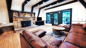 A modern living room with a stone fireplace, leather sofas, and a large window offering a view of the outdoors. - Gorgeous A Frame on 18 private acres.  (Oneonta)