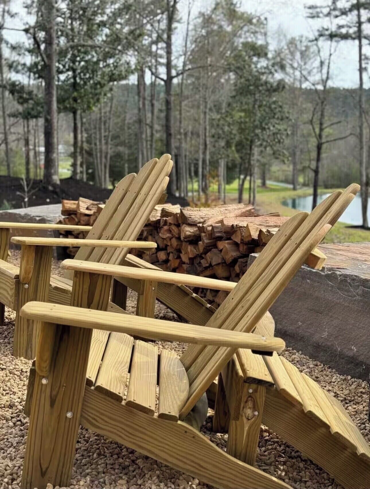 A wooden Adirondack chair with a stack of firewood behind it, set on gravel with a forest and lake in the background.