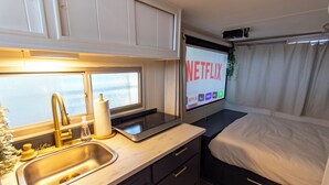 Ett kompakt sovrum med en säng, ett litet sängbord, en inbyggd tv som visar Netflix, ett fönster och ett handfat med kran.