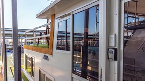 En vit byggnad med stora fönster, en träveranda och utsikt över en småbåtshamn.