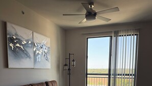 Un salón amplio con sofá de cuero marrón, ventilador de techo y un gran ventanal corredera que da a un balcón exterior.