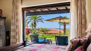 A bedroom with a large bed, a view of a garden with palm trees, and an outdoor umbrella.
