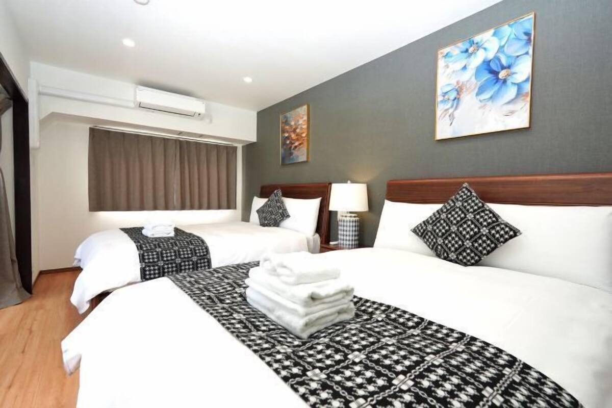 Habitación de hotel con dos camas, una cama grande con colcha estampada, una cama más pequeña con manta estampada, una mesita de noche con lámpara, aire acondicionado de pared y un cuadro.