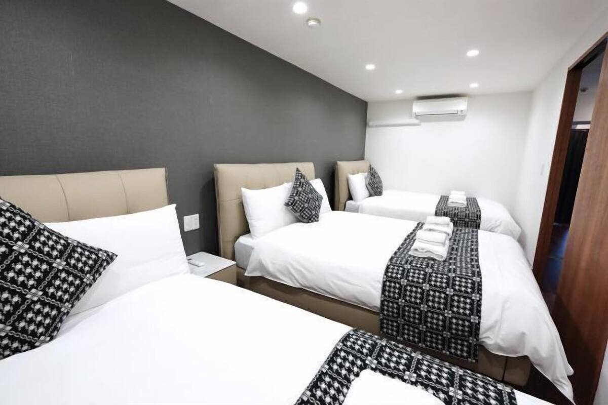 Habitación de hotel con tres camas, cada una con una colcha blanca y un plaid estampado. Una pared de color oscuro y una puerta de madera.