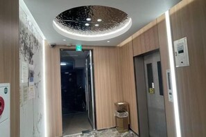 An elevator interior with a metallic finish, a wall-mounted emergency exit sign, and a door with a handle. - Nipponbashi Royal (Osaka)