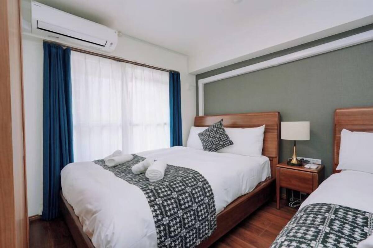 Una habitación de hotel con dos camas, un equipo de aire acondicionado, una mesita de noche con lámpara y una ventana con cortinas.