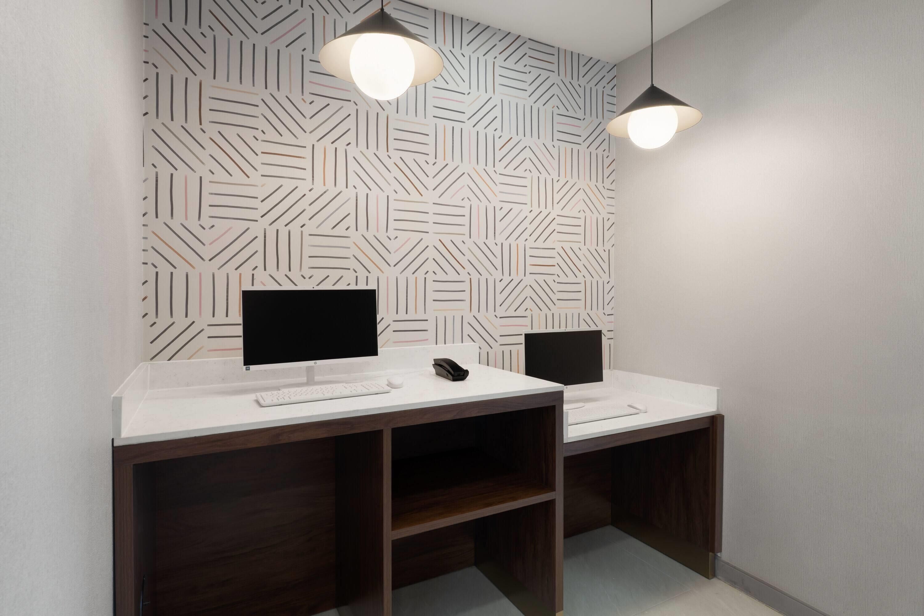 A modern office space with two desks, each with a computer monitor, keyboard, and mouse. There are two pendant lights above the desks.