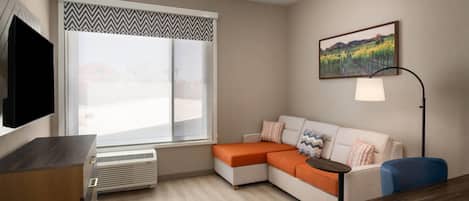 Una sala de estar moderna con un sofá modular, un televisor de pantalla plana, una lámpara de pie y un amplio ventanal con vistas a un viñedo.