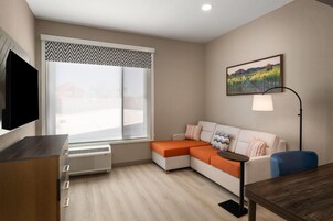 A modern living room with a sectional sofa, a flat-screen TV, a standing lamp, and a large window with a view of a vineyard.