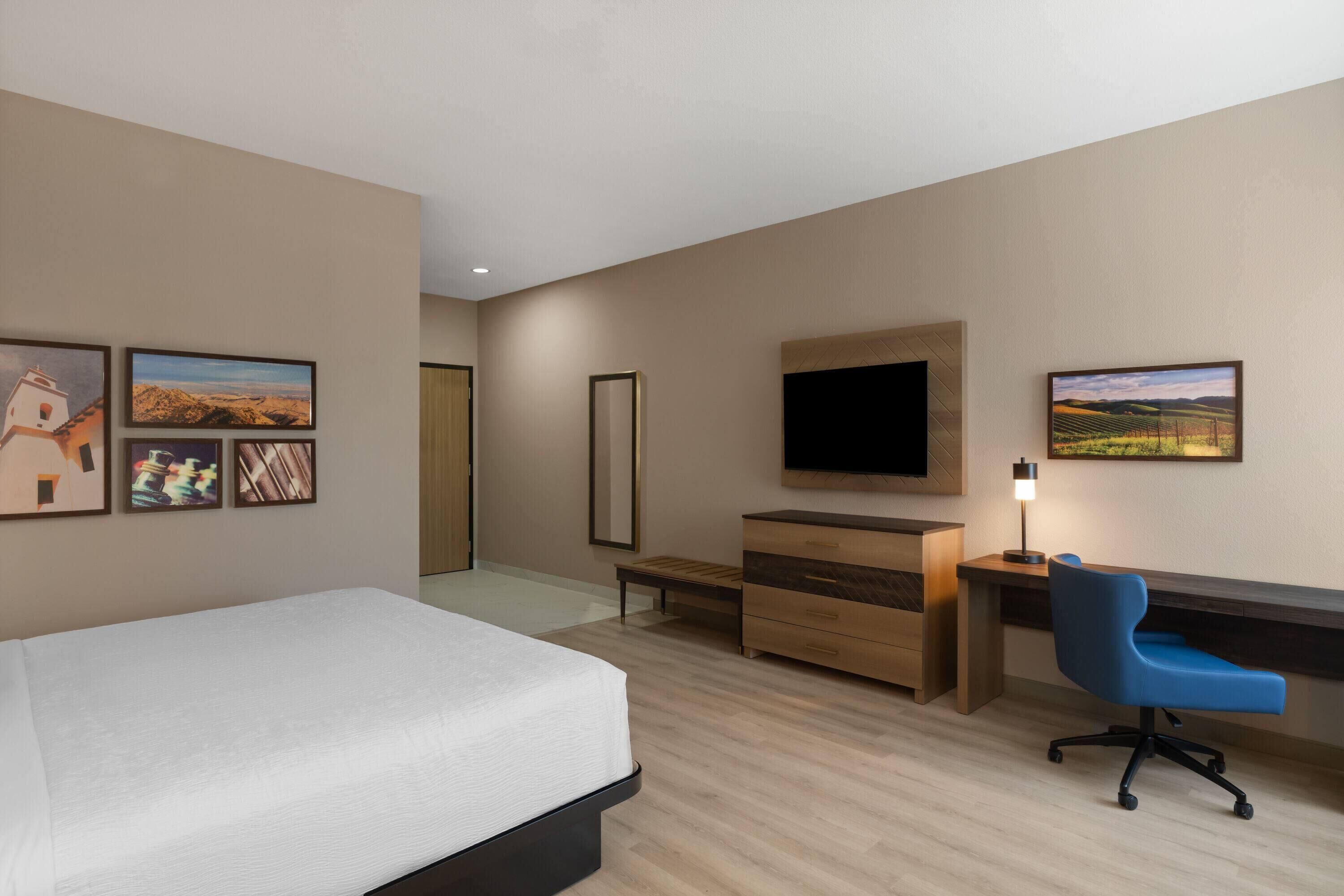 Une chambre d’hôtel équipée d’un lit, d’un bureau, d’une chaise, d’une télévision et ornée d’œuvres d’art sur les murs.