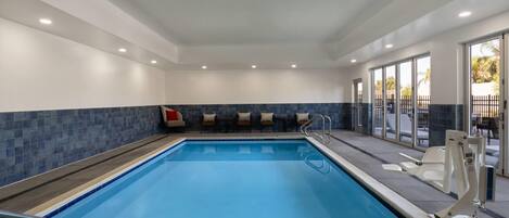 A swimming pool with a blue tiled border, a seating area with chairs and a sofa, and a glass door leading outside.