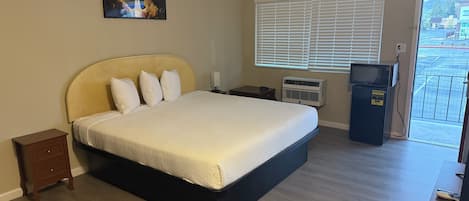A hotel room with a bed, a small bedside table, a heater, a TV, and a view of the outside.