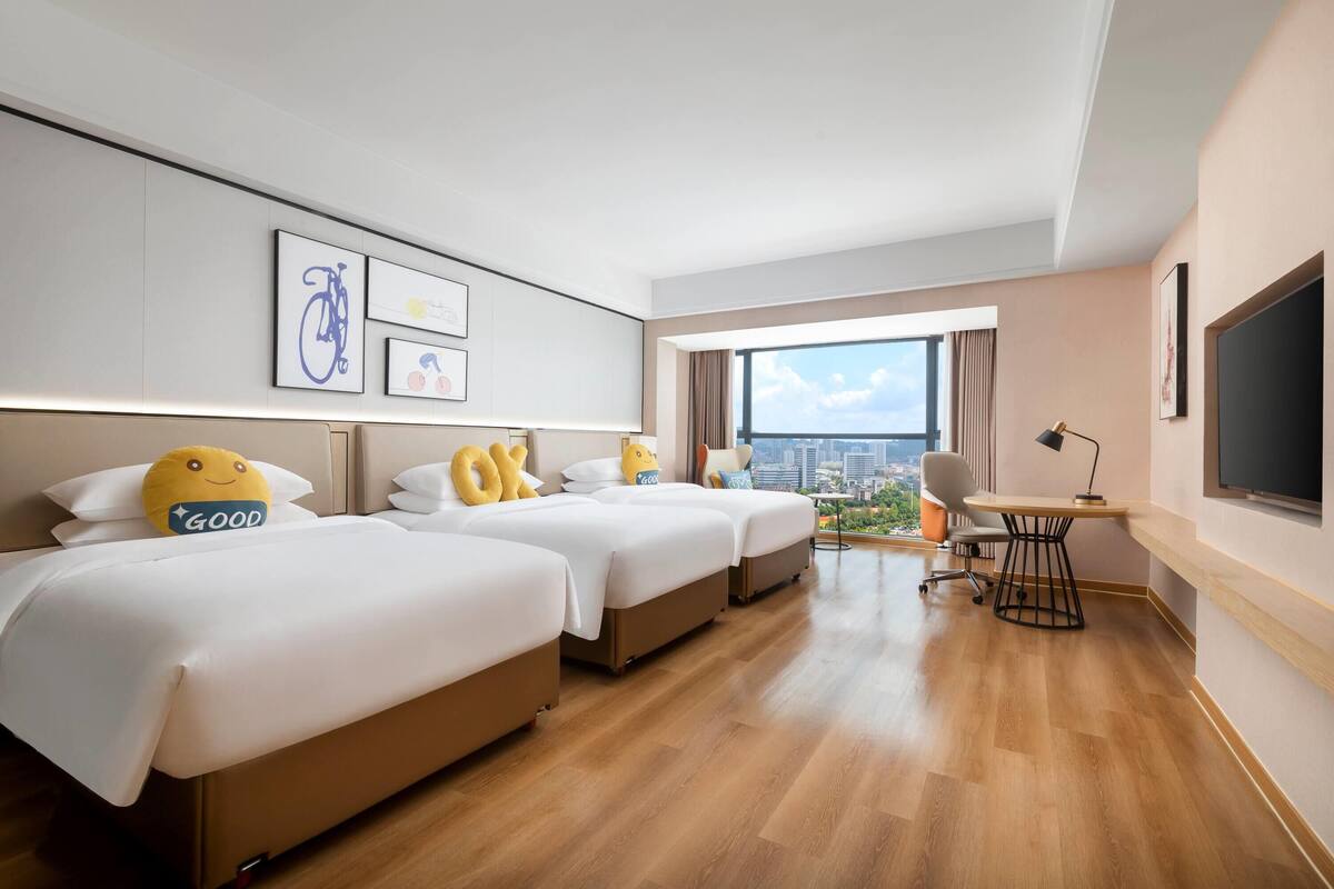 A modern hotel room with three beds, a desk, a chair, and a large window offering a city view.