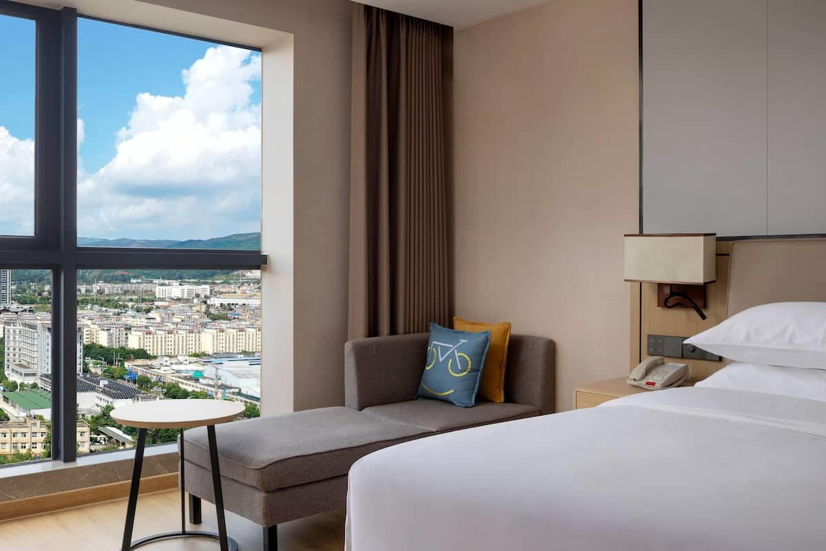 A hotel room with a large window offering a city view, a sofa with a bicycle-themed pillow, a bedside table with a phone, and a bed with white linens.