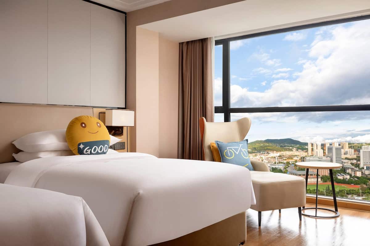 A hotel room with a large bed, a chair, a small table, and a view of a cityscape through a large window.