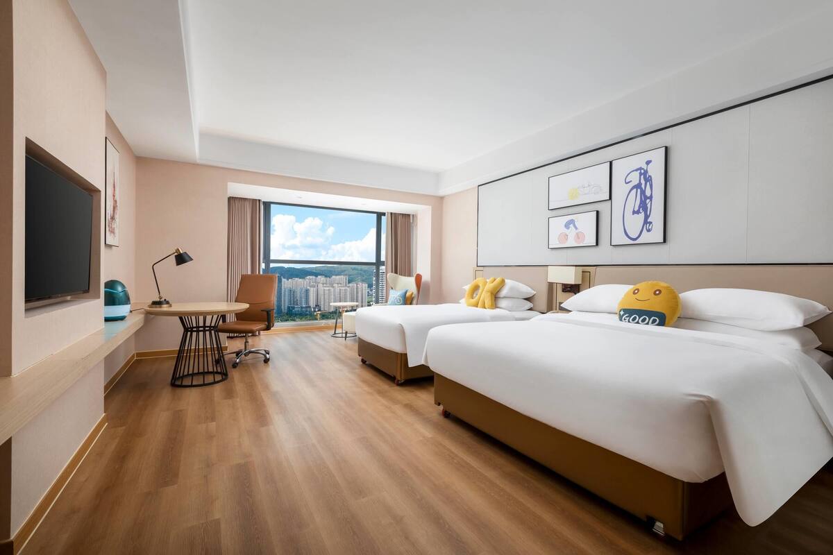 A hotel room with two beds, a desk, a chair, and a large window offering a city view.