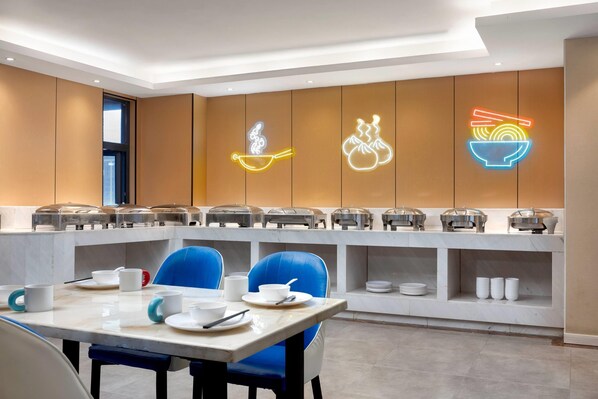 A modern breakfast area with a long counter, blue chairs, and neon signs indicating food options.