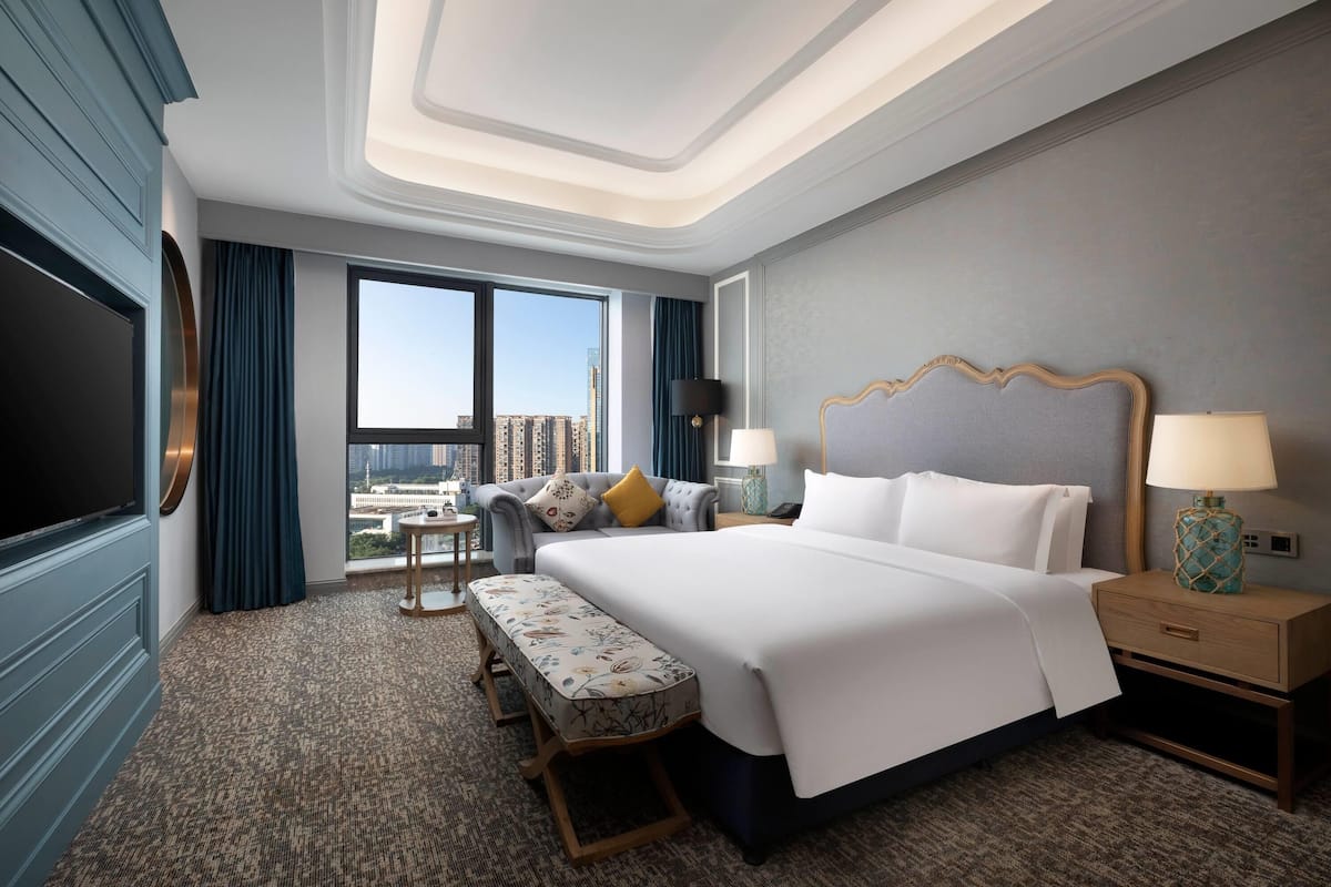 A hotel room with a large bed, a bench, a desk, a TV, and a view of the city.