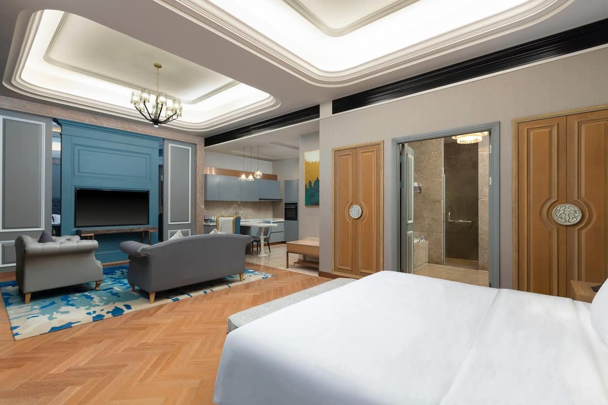 A modern hotel room with a large bed, a seating area with sofas, a TV, and a kitchenette.
