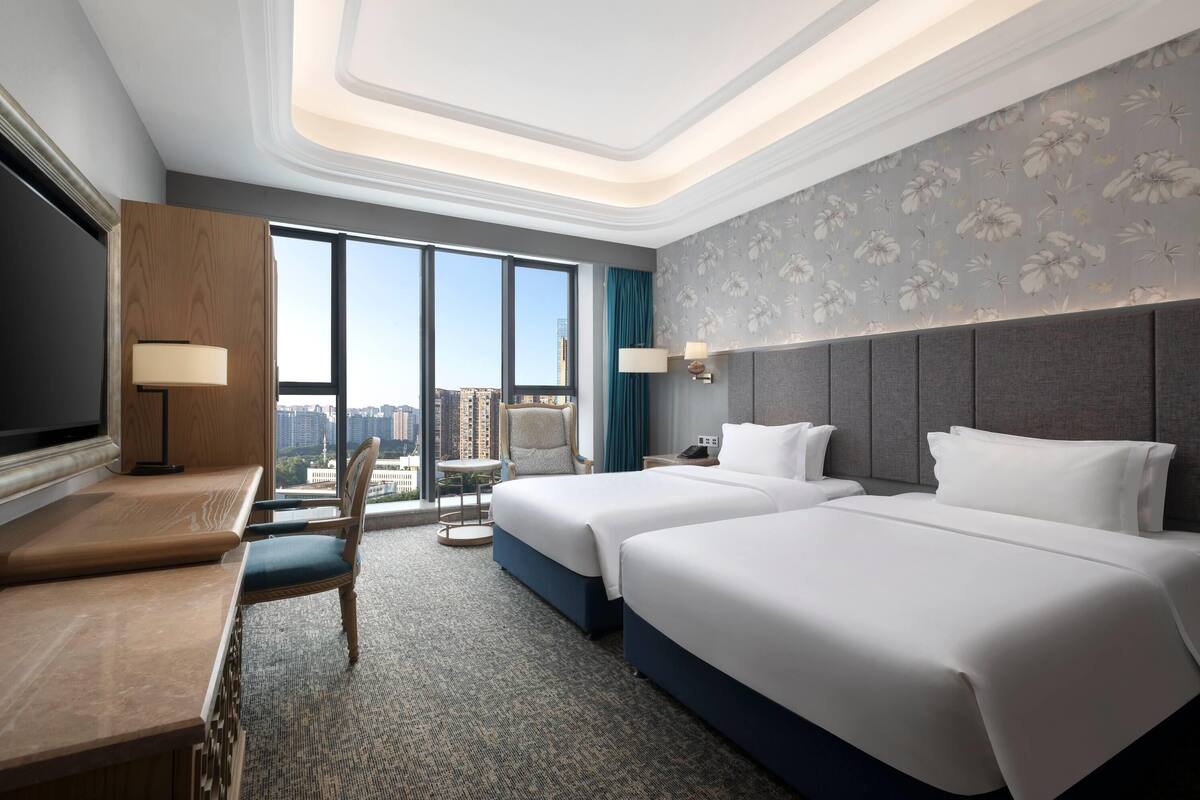 A hotel room with two beds, a desk, a chair, a TV, and a large window offering a city view.