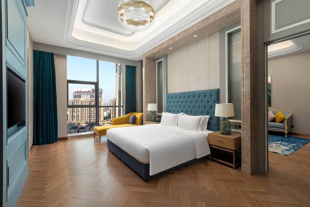 A modern hotel room with a large bed, a sofa, a TV, and a view of the city.