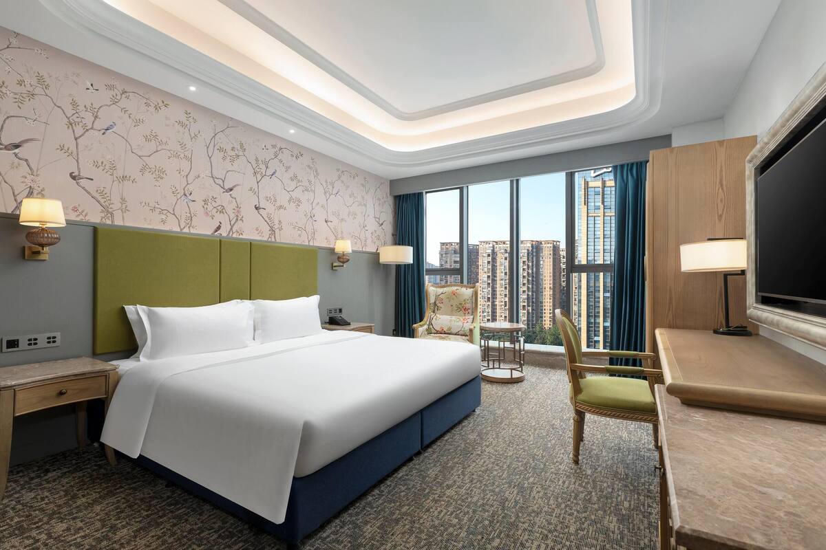 A hotel room with a large bed, a desk, a chair, a TV, and a view of tall buildings.