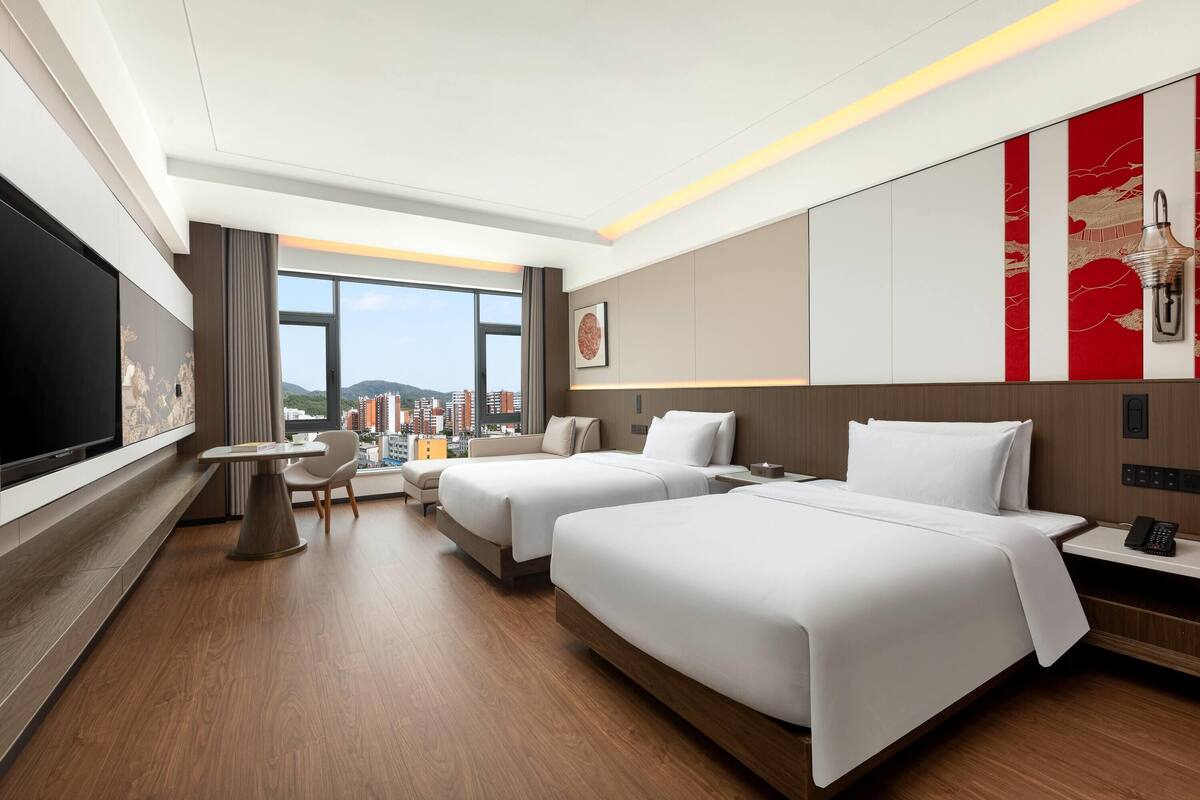 Habitación de hotel con dos camas, televisión, mesa de comedor y vista a la ciudad.