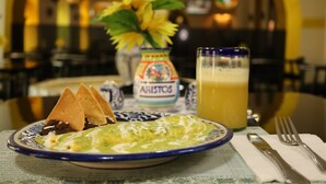 A plate with a green dish, a glass of orange juice, and a small vase with yellow flowers.
