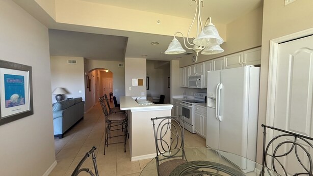 A modern kitchen with white appliances, a dining area with a glass table and metal chairs, and a hallway leading to a living room. - Cypress Woods Golf & Country Club, Beautiful 2 Bed/2Bath Condo, 1st Floor (Naples)