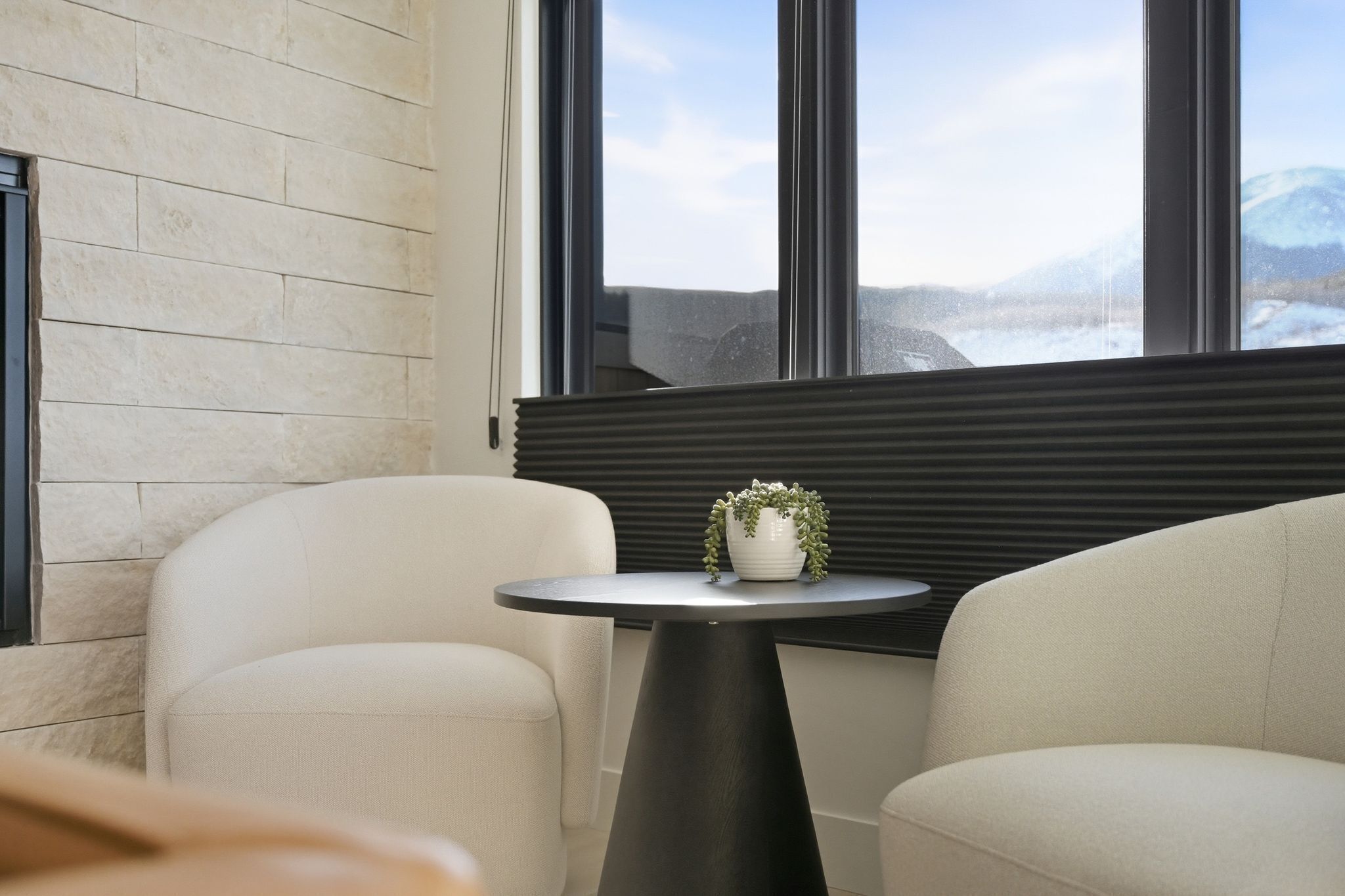 A modern room with a stone wall, two white chairs, a small round table with a plant, and a window with a view of mountains.