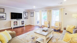 Un salon comprenant un ensemble de canapés, une table basse, un téléviseur et un meuble. Il y a des rideaux aux fenêtres et une lampe au mur.