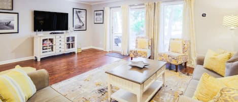 Un salon comprenant un ensemble de canapés, une table basse, un téléviseur et un meuble. Il y a des rideaux aux fenêtres et une lampe au mur.