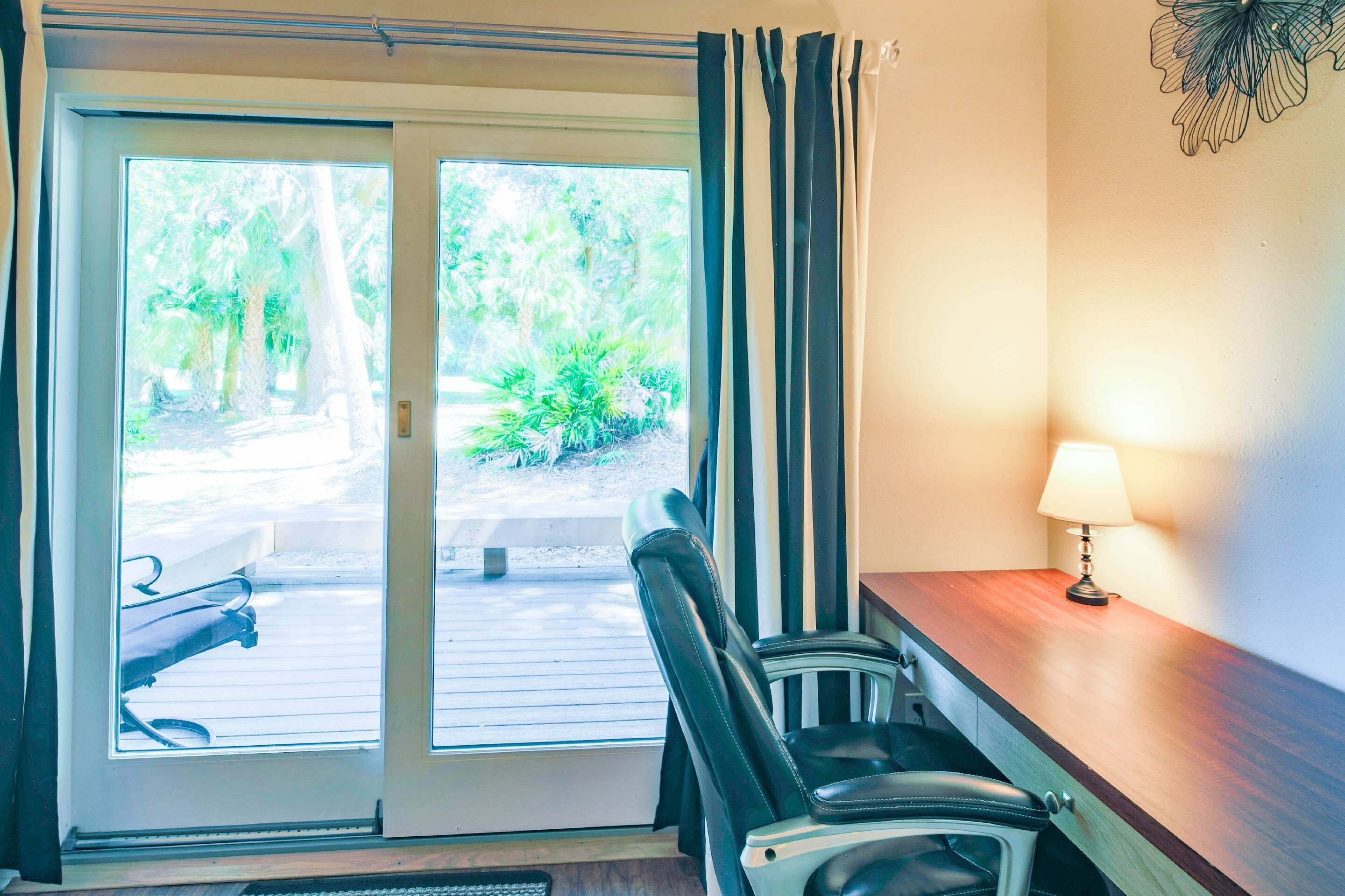 A room with a wooden desk, a chair, a lamp, and a sliding glass door leading to an outdoor area with plants.