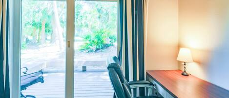 A room with a wooden desk, a chair, a lamp, and a sliding glass door leading to an outdoor area with plants.